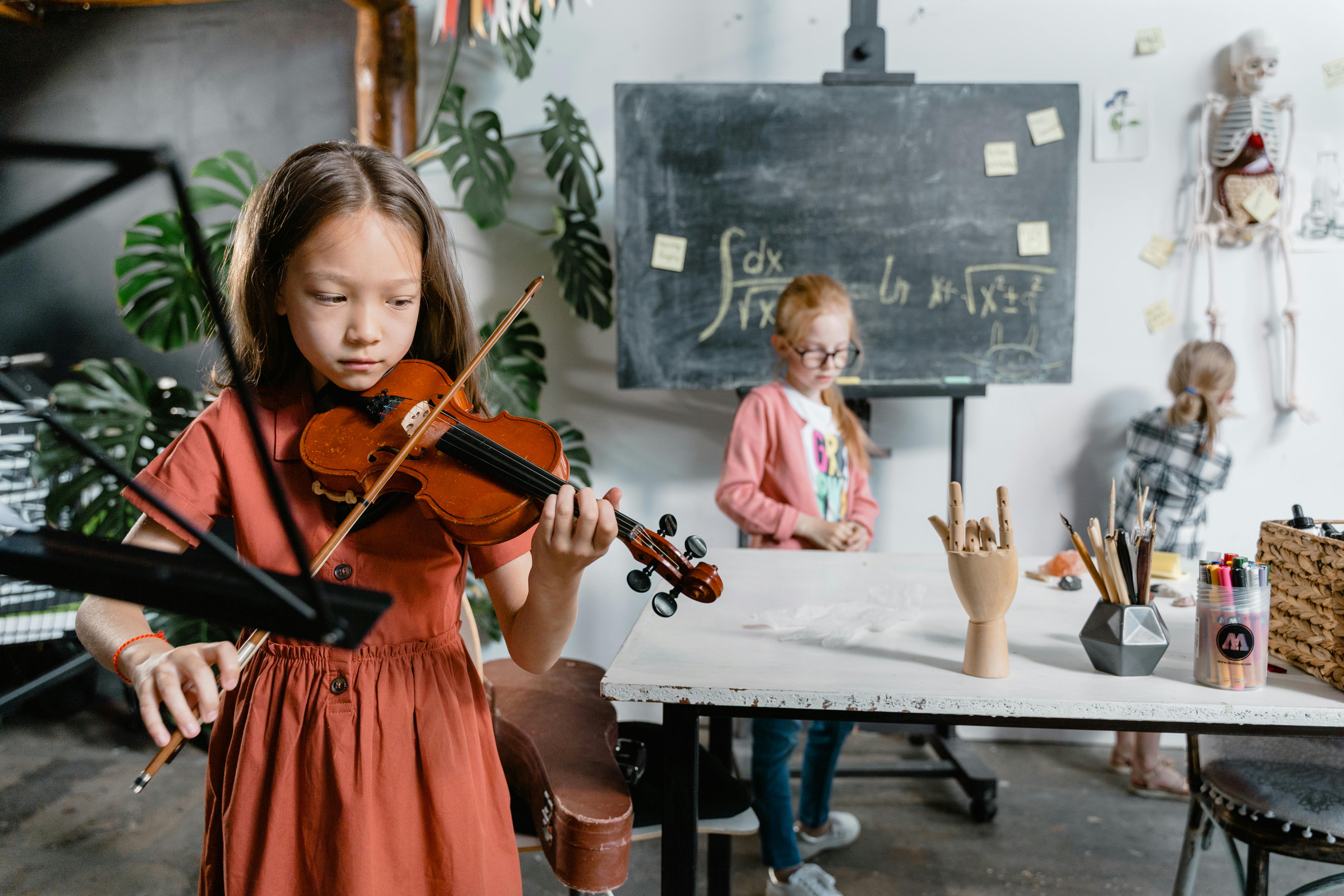Ein junges Mädchen mit einer Geige in der Hand spielt konzentriert, während andere Kinder in der Umgebung stehen oder sitzen. Die Kinder wirken wissbegierig und talentiert, die Atmosphäre ist kreativ und freundlich.
